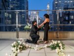 Man proposing on one knee on an elevated rooftop terrace with white floral heart arrangement and sweeping Manhattan skyline views at twilight
