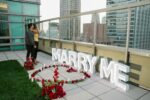 Overhead view of MARRY ME letter signs encircled by red rose petals on a rooftop terrace with the New York City skyline behind
