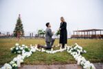 Couple sharing a proposal moment in a flower-lined heart arrangement on a lawn with a Christmas tree and NYC waterfront in winter