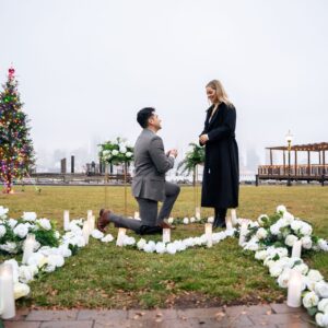 Couple sharing a proposal moment in a flower-lined heart arrangement on a lawn with a Christmas tree and NYC waterfront in winter
