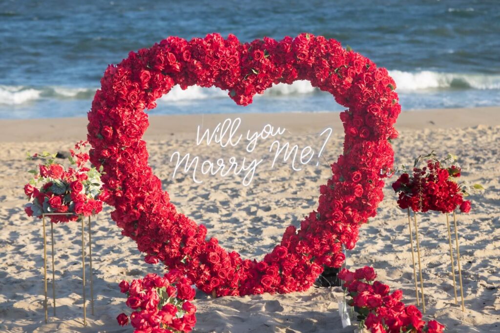 Red rose heart arch with 'Will You Marry Me' script on a sandy beach at the ocean's edge, with candles and rose petals surrounding the display