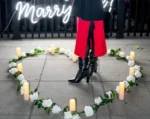 Woman standing in a candle-lit rose heart arrangement with a 'Will You Marry Me' neon sign against the glittering New York City waterfront at night
