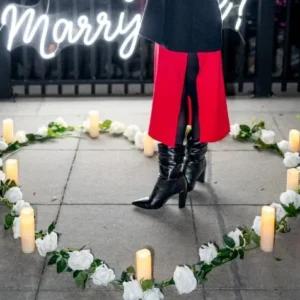 Woman standing in a candle-lit rose heart arrangement with a 'Will You Marry Me' neon sign against the glittering New York City waterfront at night